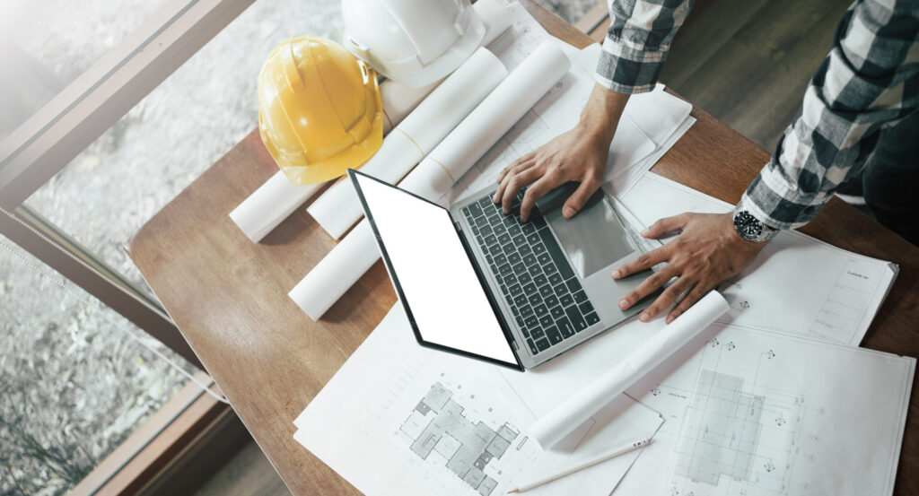 Close-up of asian male civil engineer working with drawings inspection on laptop on construction site at work desk in office.