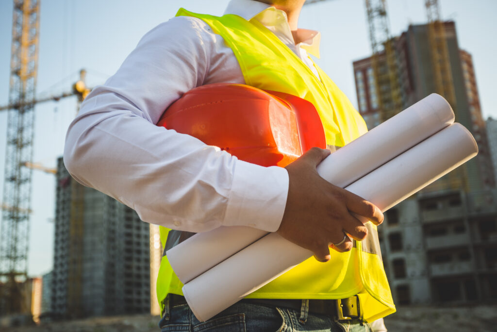 Closeup photo of man in shirt and jacket holding hardhat and blueprints on building site