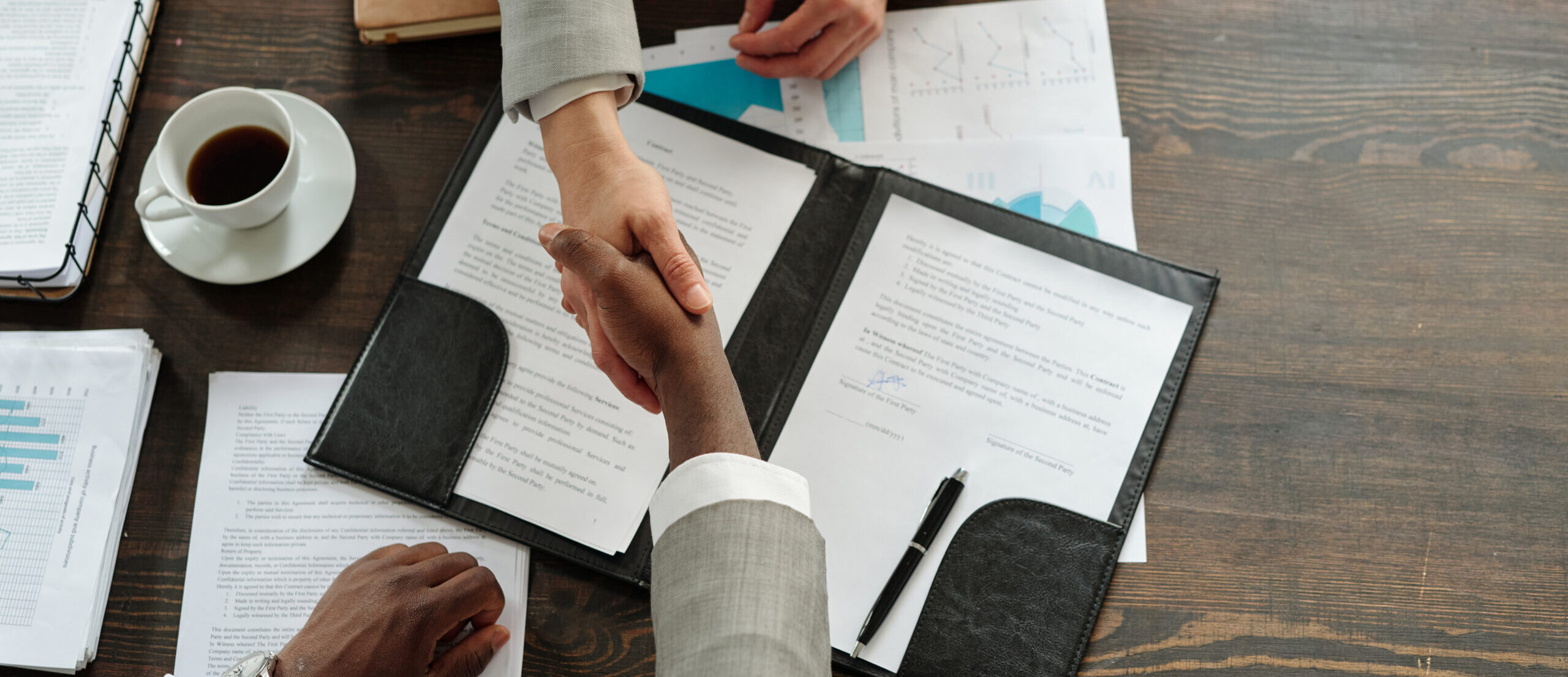 Caucasian woman and Black man shaking hands over contract documents on wooden desk, both wearing business attire, coffee cup and financial charts visible, top view composition