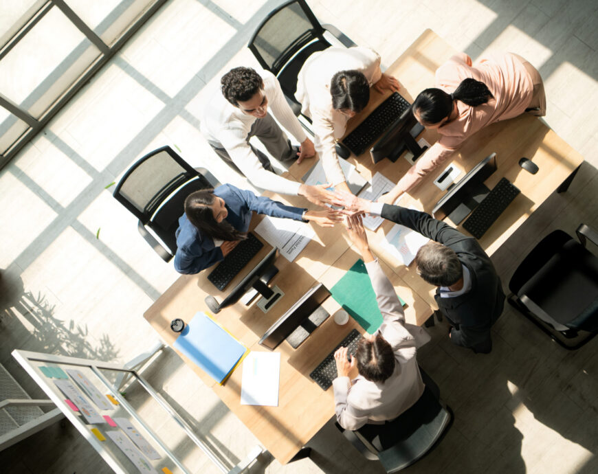 Top view of a multicultural business team stacking hands together over a meeting table, symbolizing unity, teamwork, collaboration, trust, and shared goals in a professional office setting