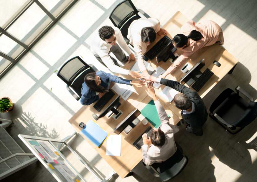 Top view of a multicultural business team stacking hands together over a meeting table, symbolizing unity, teamwork, collaboration, trust, and shared goals in a professional office setting